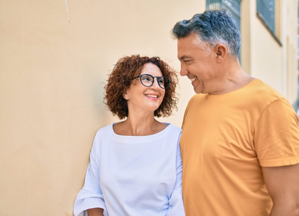A smiling couple stands close together against a light-colored wall, gazing at each other with warmth and affection. The woman wears glasses and a white top, while the man wears a mustard-yellow shirt, both appearing happy and relaxed in a sunlit setting.