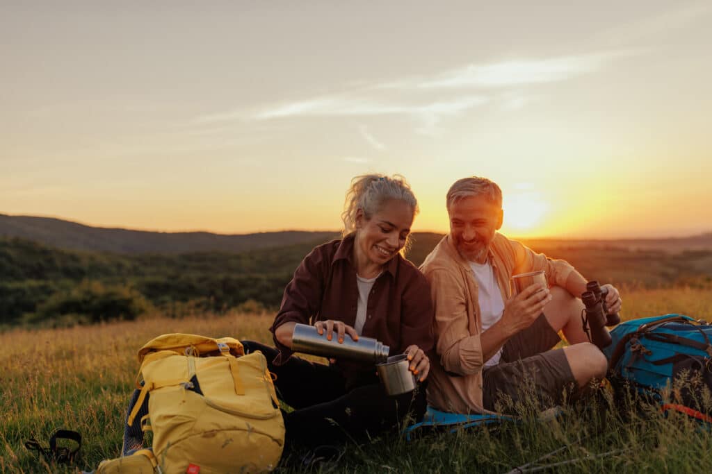 A smiling couple sits in a grassy field at sunset, enjoying hot drinks with camping gear and backpacks beside them. The woman pours from a thermos while the man holds a cup and binoculars, both relaxed and soaking in the scenic view.