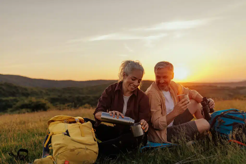 A smiling couple sits in a grassy field at sunset, enjoying hot drinks with camping gear and backpacks beside them. The woman pours from a thermos while the man holds a cup and binoculars, both relaxed and soaking in the scenic view.
