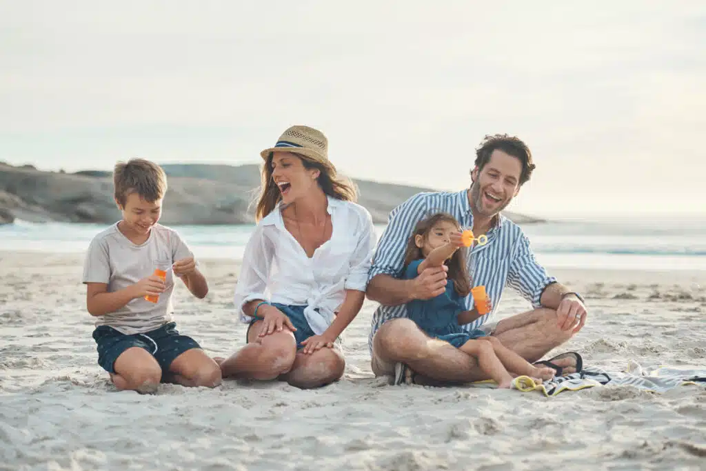 A family of four sits together on the sand at the beach, smiling and laughing as the children play with bubble wands. The parents appear relaxed and joyful, enjoying the sunshine and ocean breeze with a calm shoreline in the background.