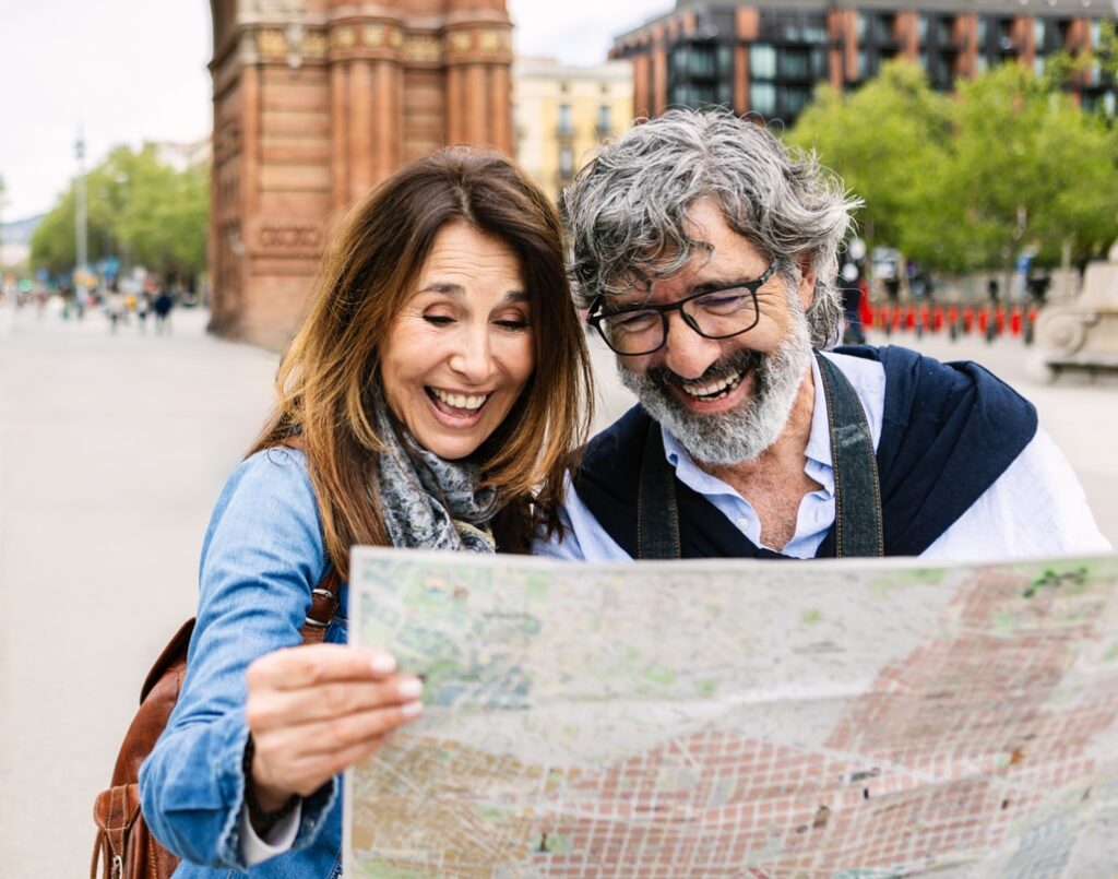 A joyful couple stands outdoors in a city plaza, smiling and laughing as they look at a large map. With a historic archway and modern buildings in the background, the scene suggests they're exploring a vibrant, culturally rich destination—likely on a travel adventure together.