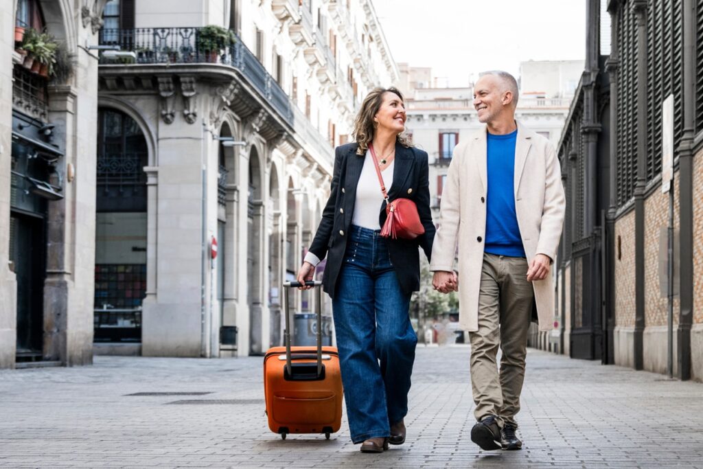 A stylish couple walks hand-in-hand through a European-style city street, smiling at each other. The woman pulls a bright orange suitcase, suggesting they’re either arriving or departing for a trip. The elegant architecture around them enhances the feel of an urban getaway filled with charm and connection.