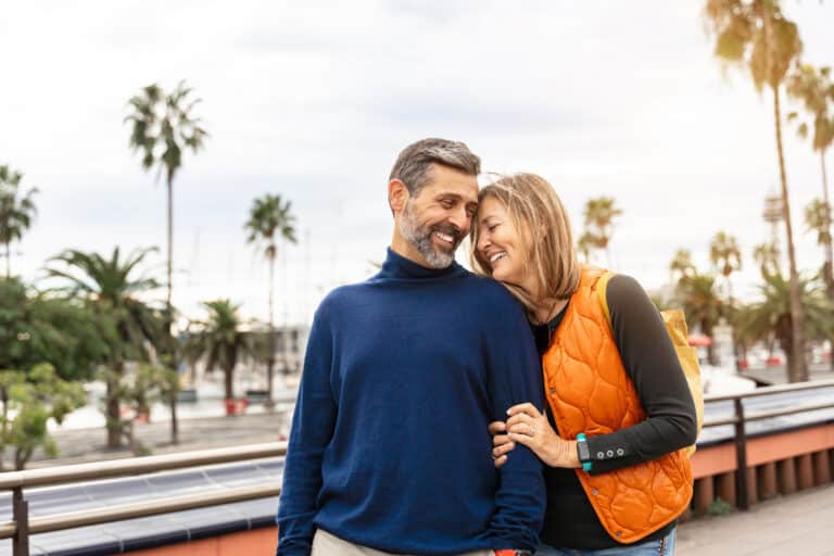 A couple stands close together outdoors, smiling with their foreheads touching. The man wears a navy sweater while the woman leans on him in an orange vest, with palm trees and a marina in the background.